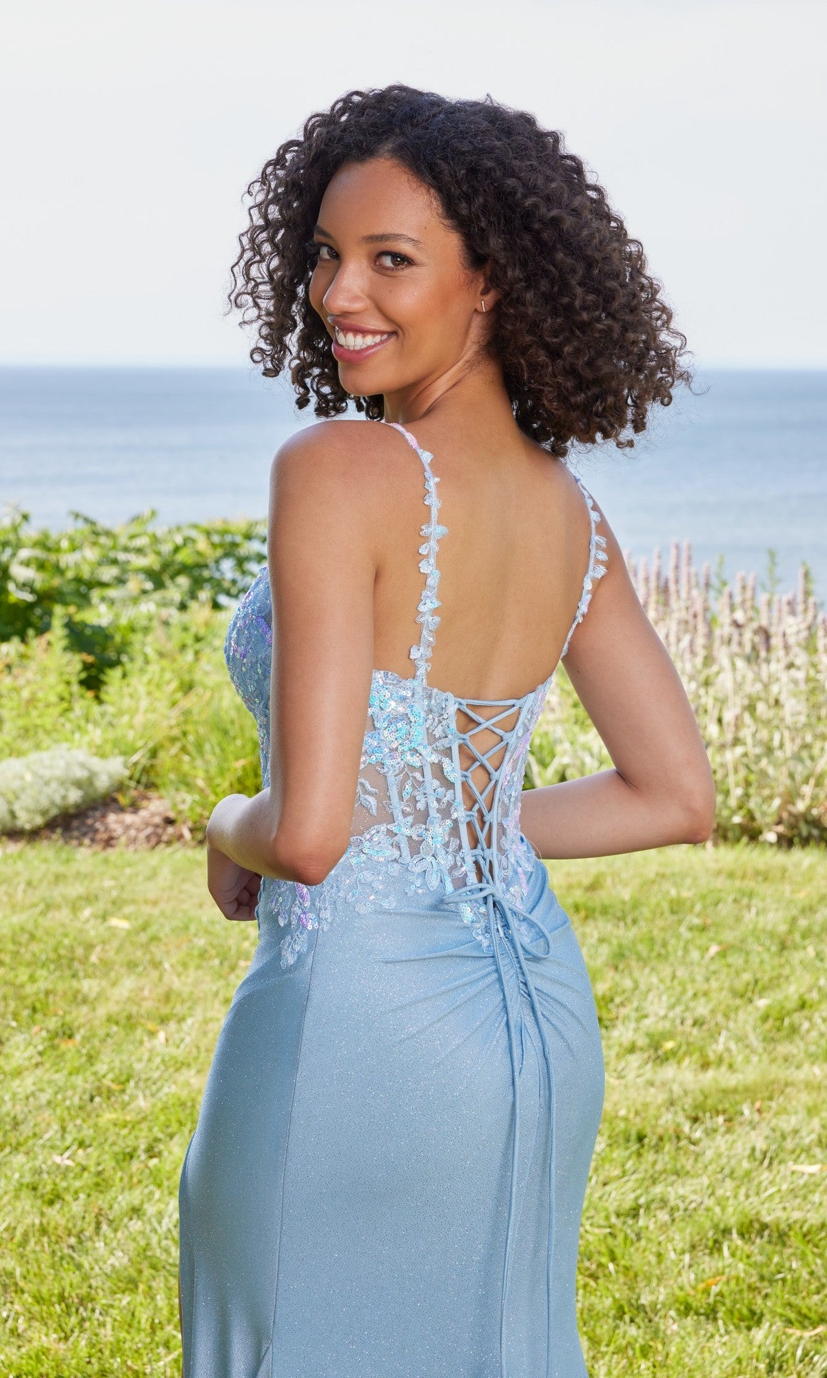 A woman with curly hair smiles outdoors on grass, wearing the Long Prom Dress: Morilee 49132 in light blue, with the ocean and greenery visible in the background.